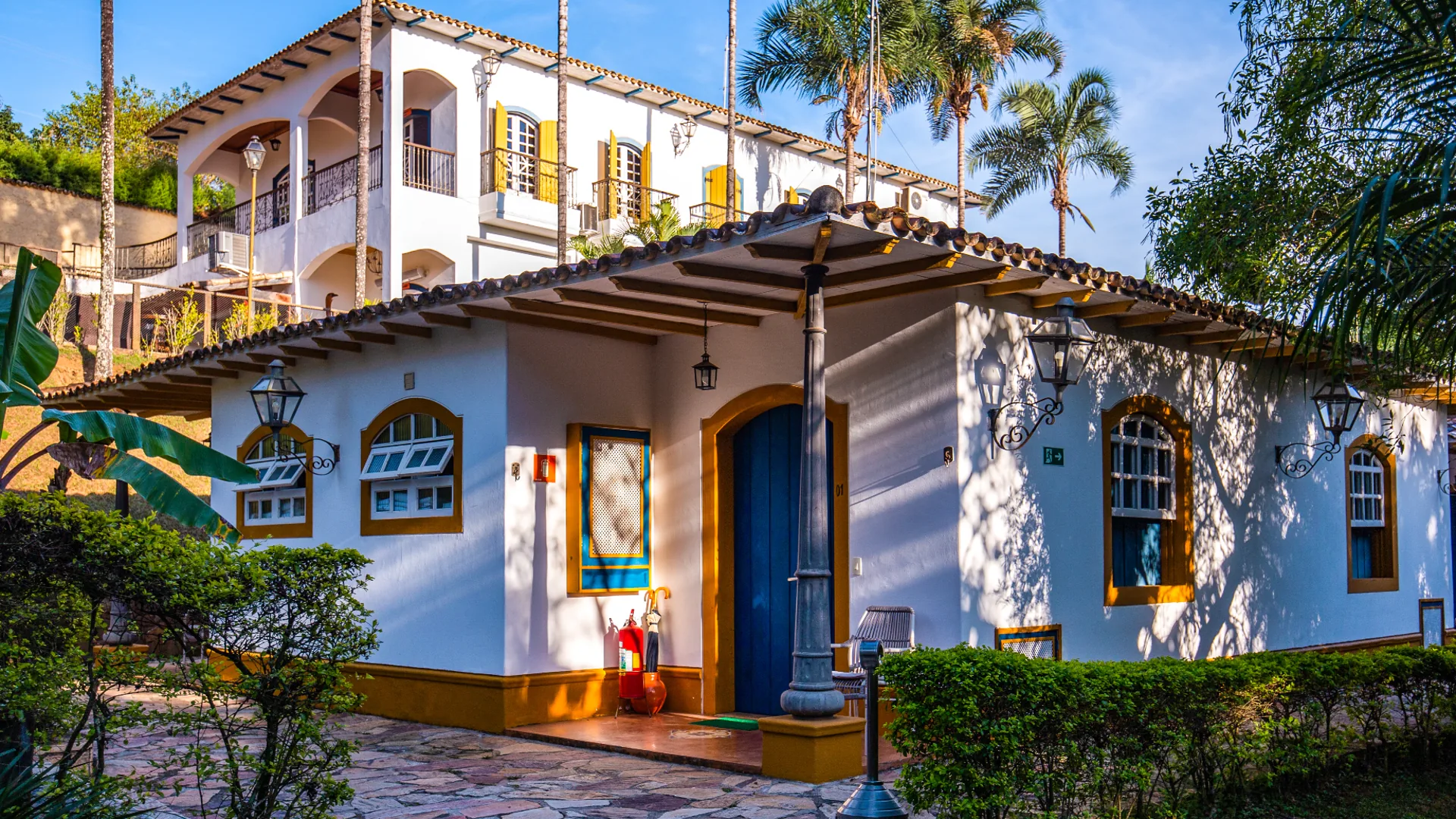 Fachada branca da Confraria Colonial, com janelas azuis e varanda voltada para a mata.